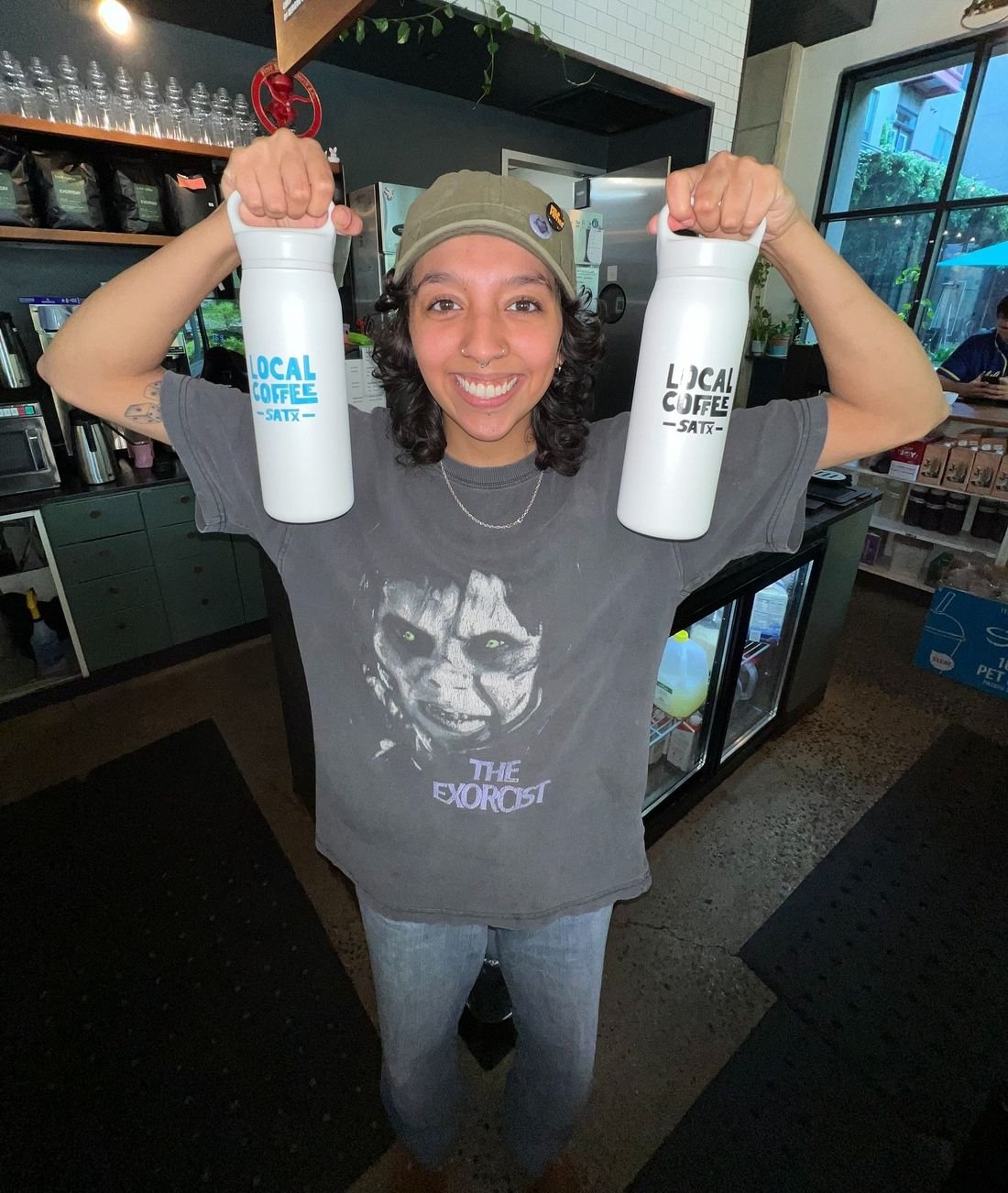 A smiling Local Coffee barista holding up two Local Coffee SATX bottles
